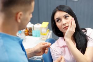 Woman with black hair in a pink shirt sitting in dentist's chair looking at the dentist while holding her hand to her jaw