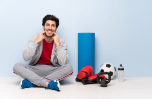 Man in gray sweats and a red shirt smiling and pointing to his teeth sitting next to athletic equipment