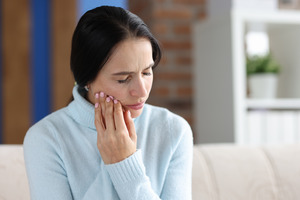 Woman in sweater sitting on couch rubbing jaw in pain